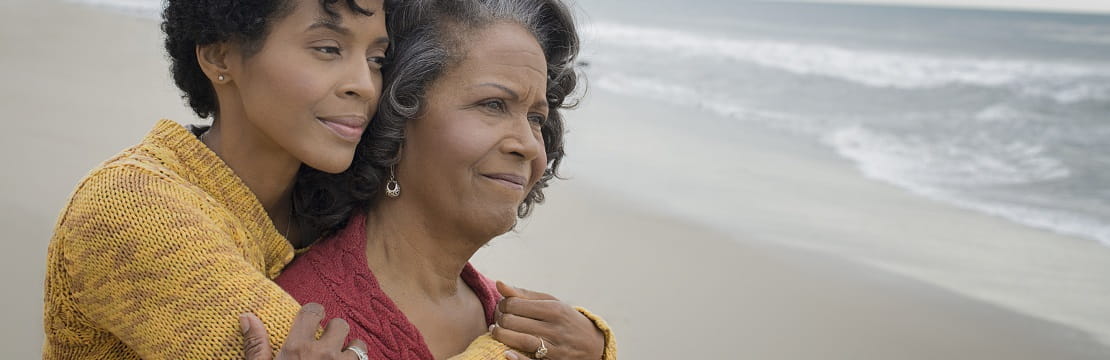 Woman hugs her mother from behind as they look out over the beach.